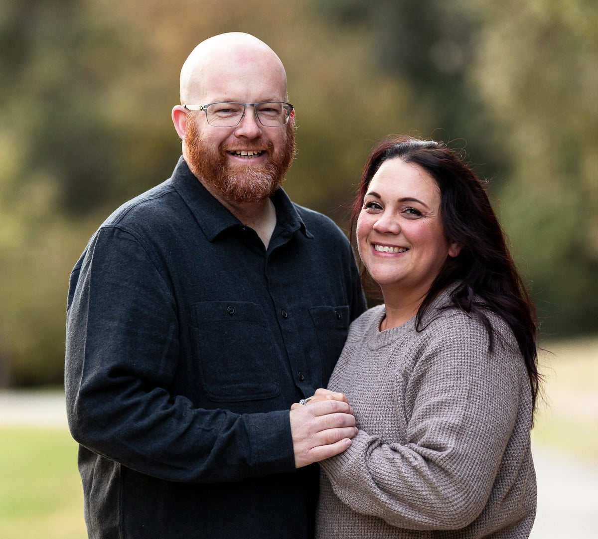 A smiling bald man with a beard and glasses wearing a dark button-up shirt, standing next to a smiling woman with long dark hair wearing a textured taupe knit sweater, set against a blurred outdoor background of trees and grass.